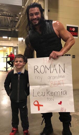 Roman Reigns and a kid posing for a photo. Roman is holding a poster that says: "Hey ROMAN my gandma just BEAT Leukemia too!"